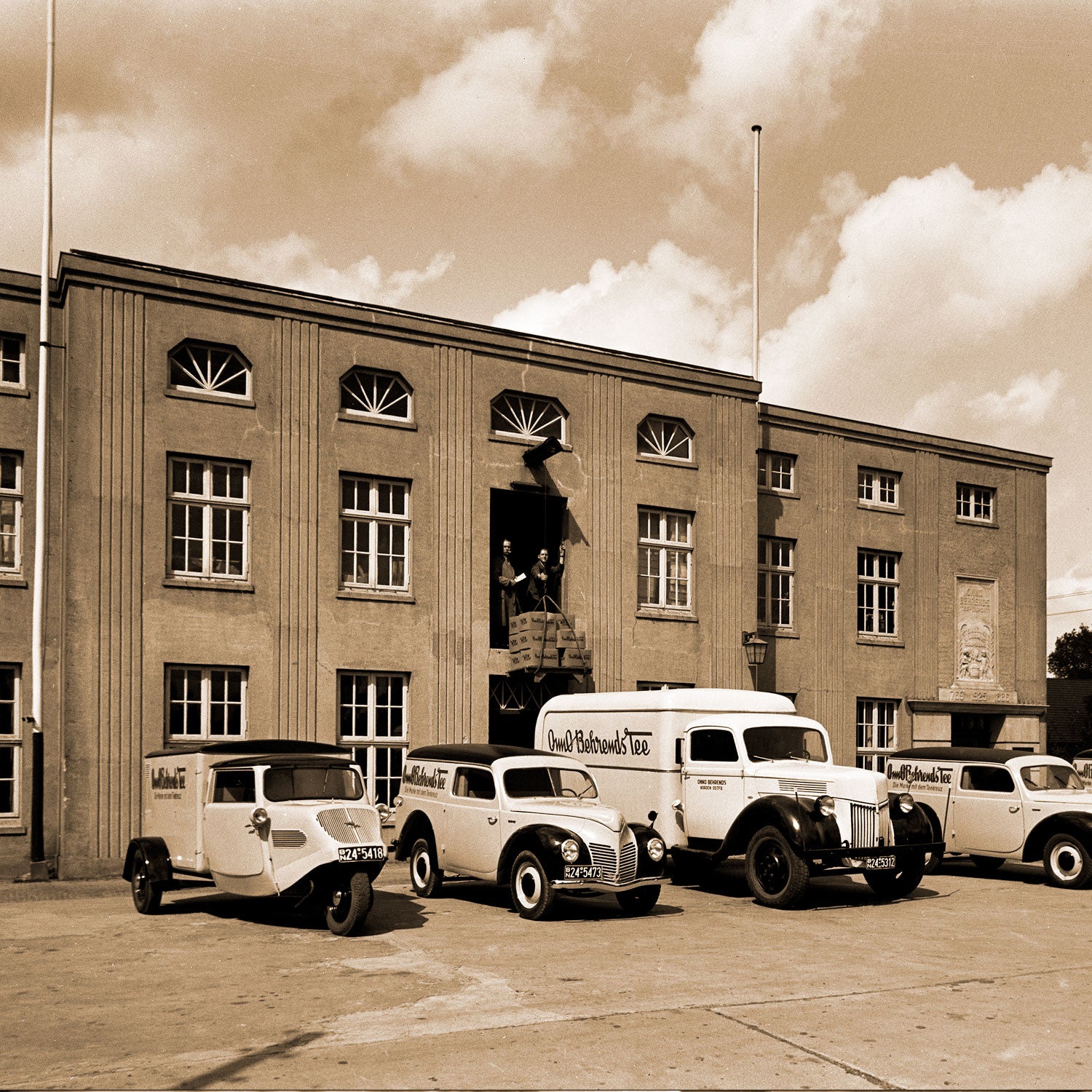 Altes Schwarz-Weiß Foto mit 4 historischen Lieferwagen von Onno Behrends Tee vor dem Betriebsgebäude.