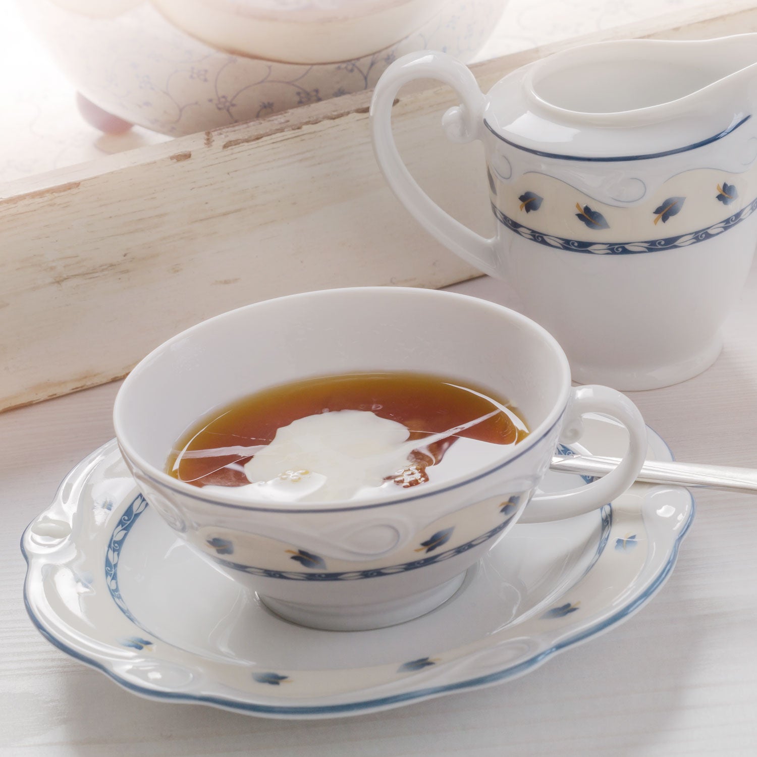 Teetasse gefüllt mit Schwarztee und einem Milchwölkchen, im Hintergrund ein Milchkännchen.