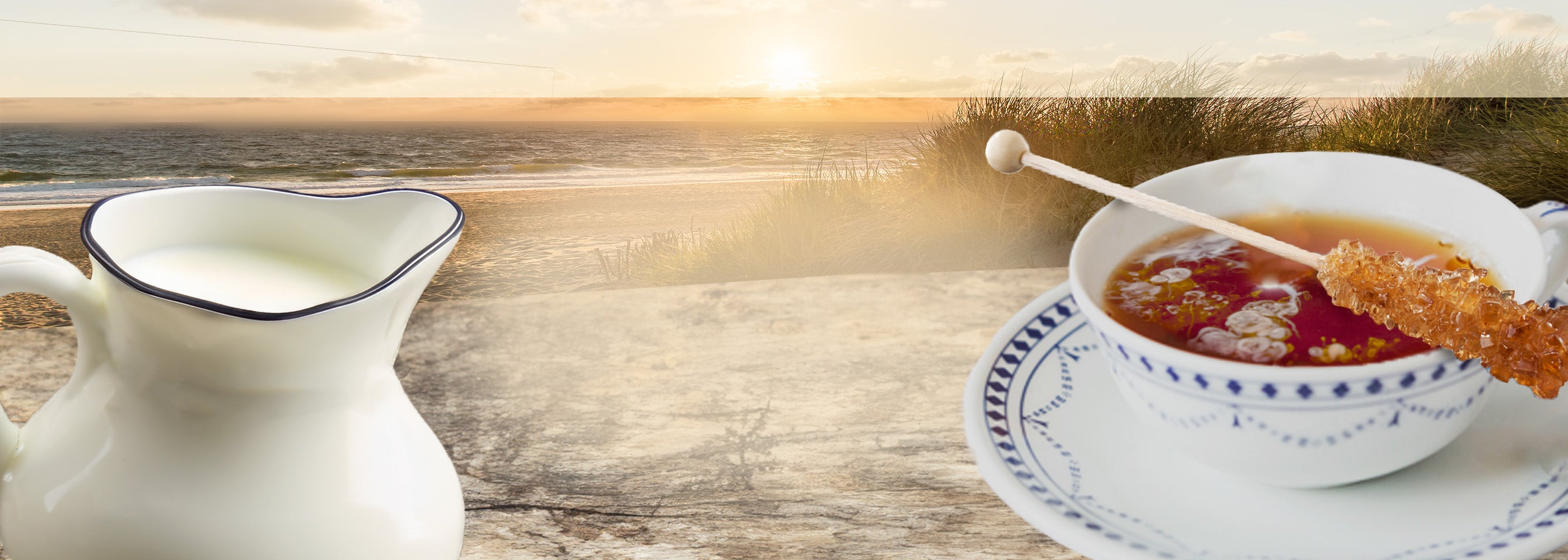 Im Vordergrund eine Tasse Schwarztee mit aufliegendem Kandiszucker, daneben ein Milchkännchen und im Hintergrund ein Nordseestrand mit untergehender Sonne.