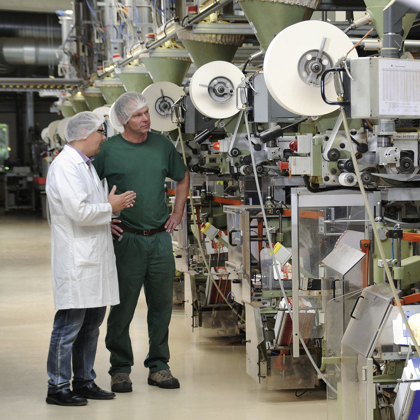 2 Mitarbeiter reden miteinander vor einer Reihe Verpackungsmaschine in der Produktionshalle von Onno Behrends.